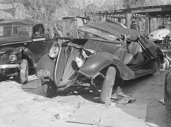 A car with its roof and body crushed, standing in a yard beside another damaged car, 1940